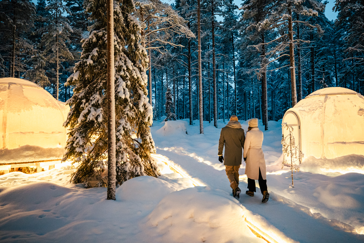 Couple walking hand in hand between glowing glass igloos in the snow at Invisible Forest Lodge.
