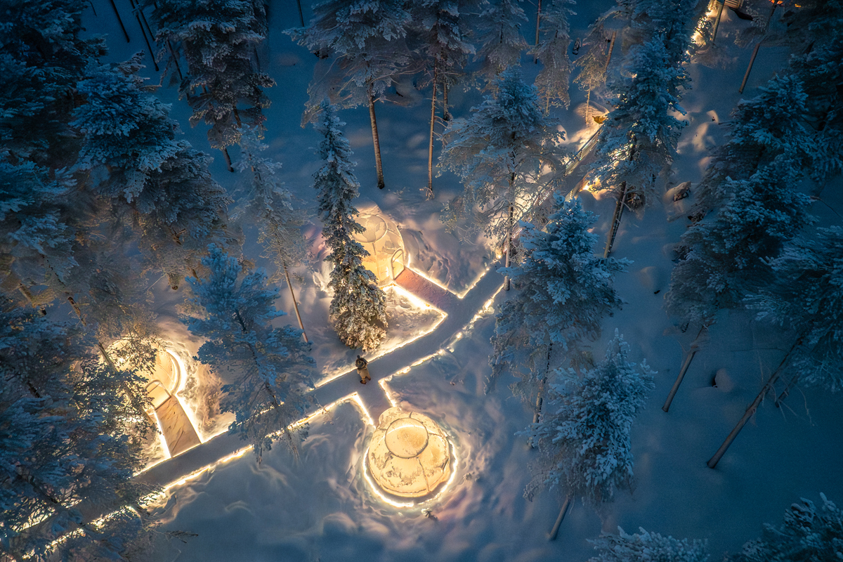 Aerial night view of illuminated glass igloos connected by lit pathways in a snowy forest at Invisible Forest Lodge.