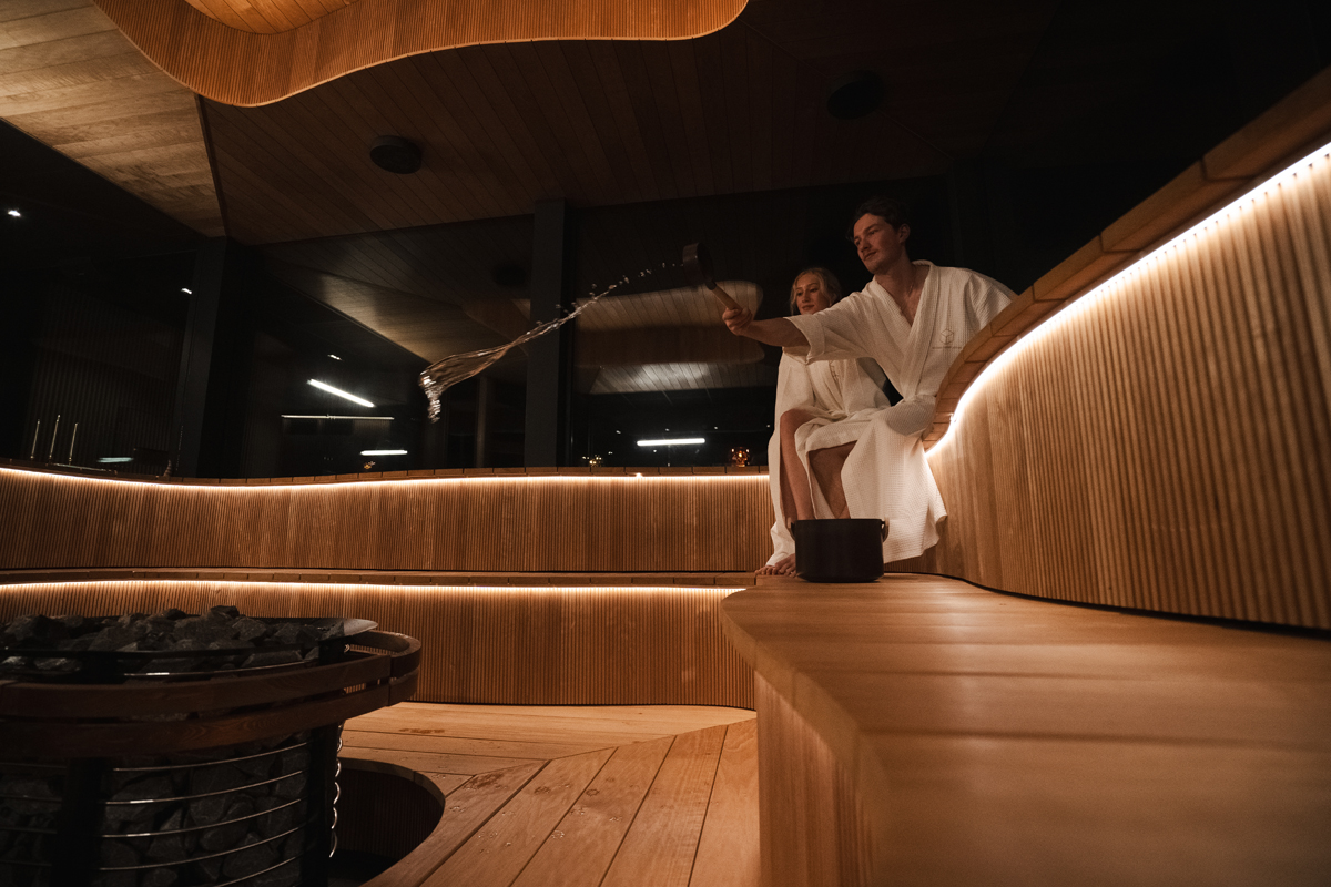 Guests in white robes throwing water on hot sauna stones inside the curved wooden sauna at Invisible Forest Lodge.