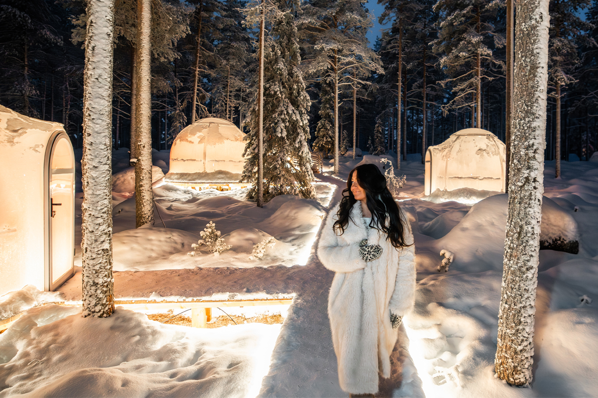 Woman in a white fur coat walking among illuminated glass igloos in a snowy forest at Invisible Forest Lodge.