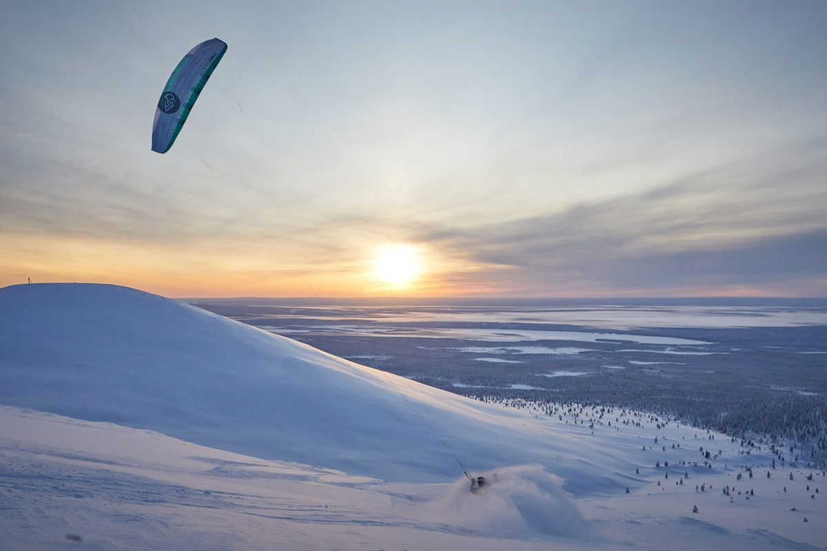 A snow kiter descends a steep snowy slope at sunset over a wide Lapland landscape during a snow kiting session.