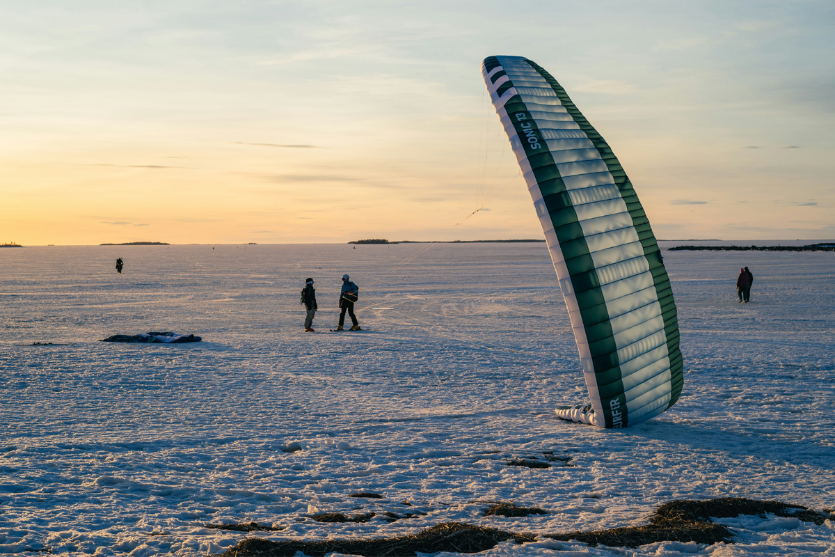 A large snow kite stands upright on a frozen lake at sunset while two kiters prepare nearby in Lapland.