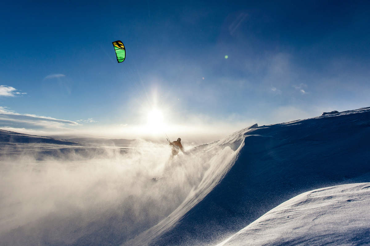 A snow kiter cuts through powder snow on a bright Lapland hillside, a green kite overhead against a deep blue sky.
