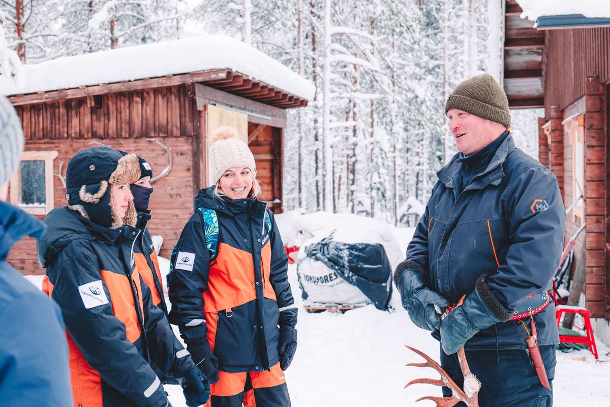 A group in winter gear talks with a local guide holding reindeer antlers outside a wooden cabin.