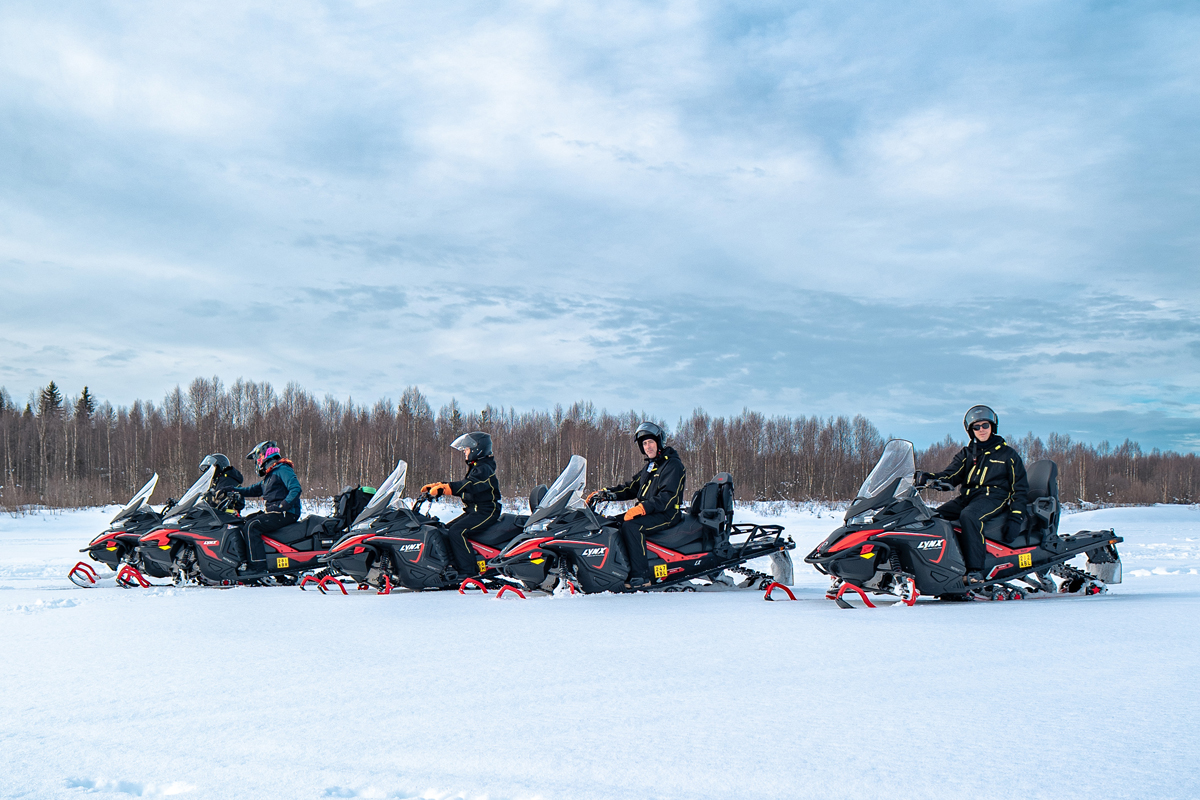 A row of five snowmobiles with riders pauses on an open snowfield near a forest edge in Lapland.