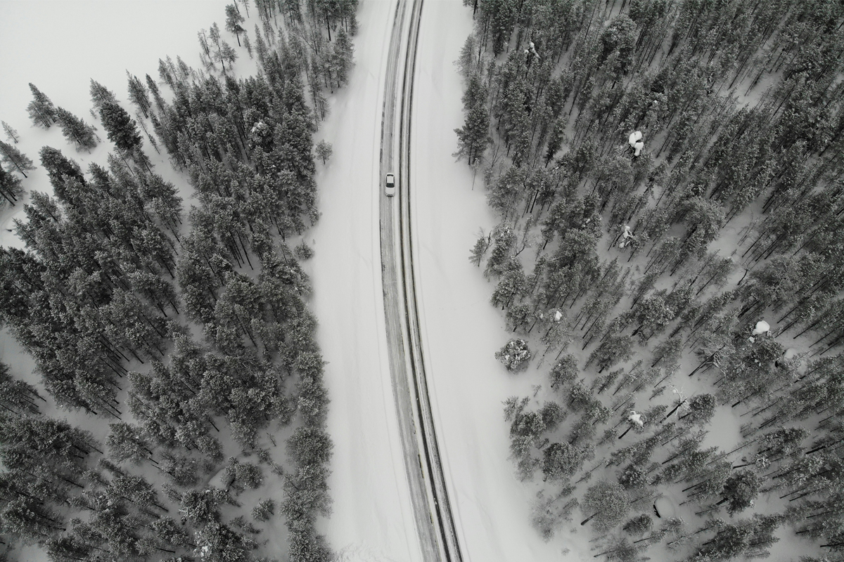 Aerial view of a single car driving a snow-covered road through dense pine forest in Lapland.
