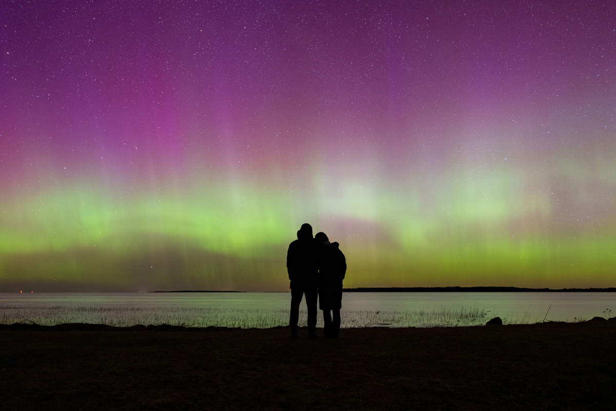 Silhouetted couple watching green and purple Northern Lights over a lake at night