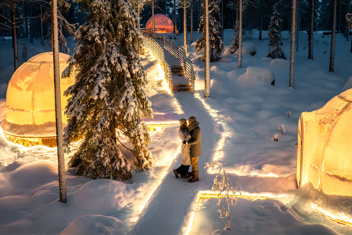Couple embracing on a lit snowy path between glass igloos at Invisible Forest Lodge
