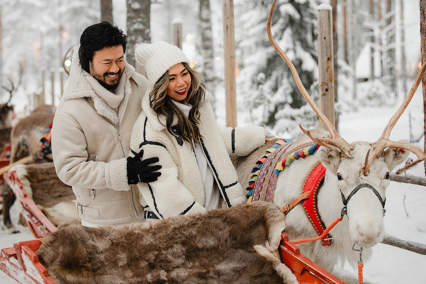 Smiling couple beside a reindeer and sleigh in a snowy forest in Lapland