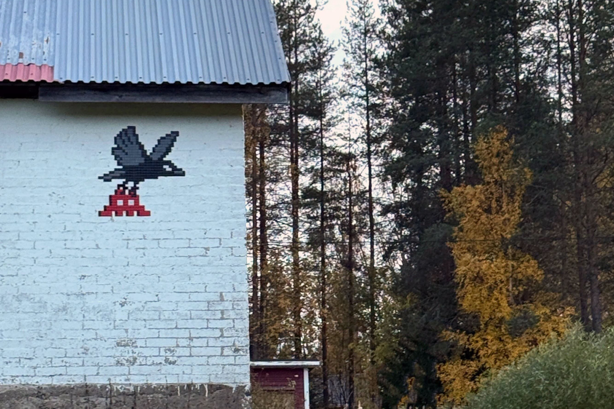 Pixel mosaic of a black crow perched on a red invader on a white wall beside autumn forest ©Invader @invaderwashere
