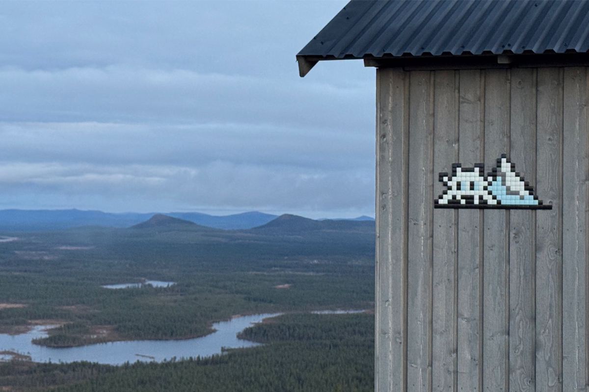 Pixel mosaic of an ice-blue invader on a wooden wall overlooking lakes and fells in Lapland ©Invader @invaderwashere