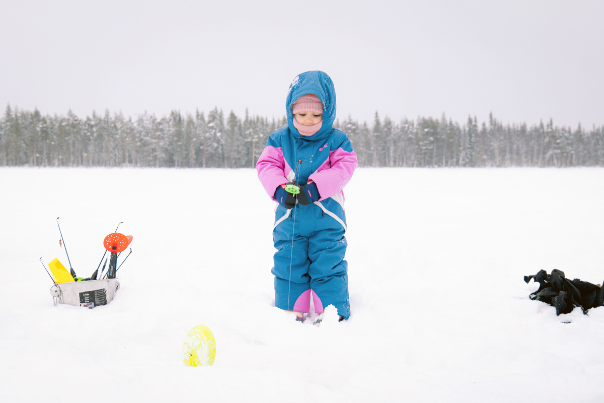 Toddler in a blue snowsuit ice fishing on a frozen lake in Finnish Lapland
