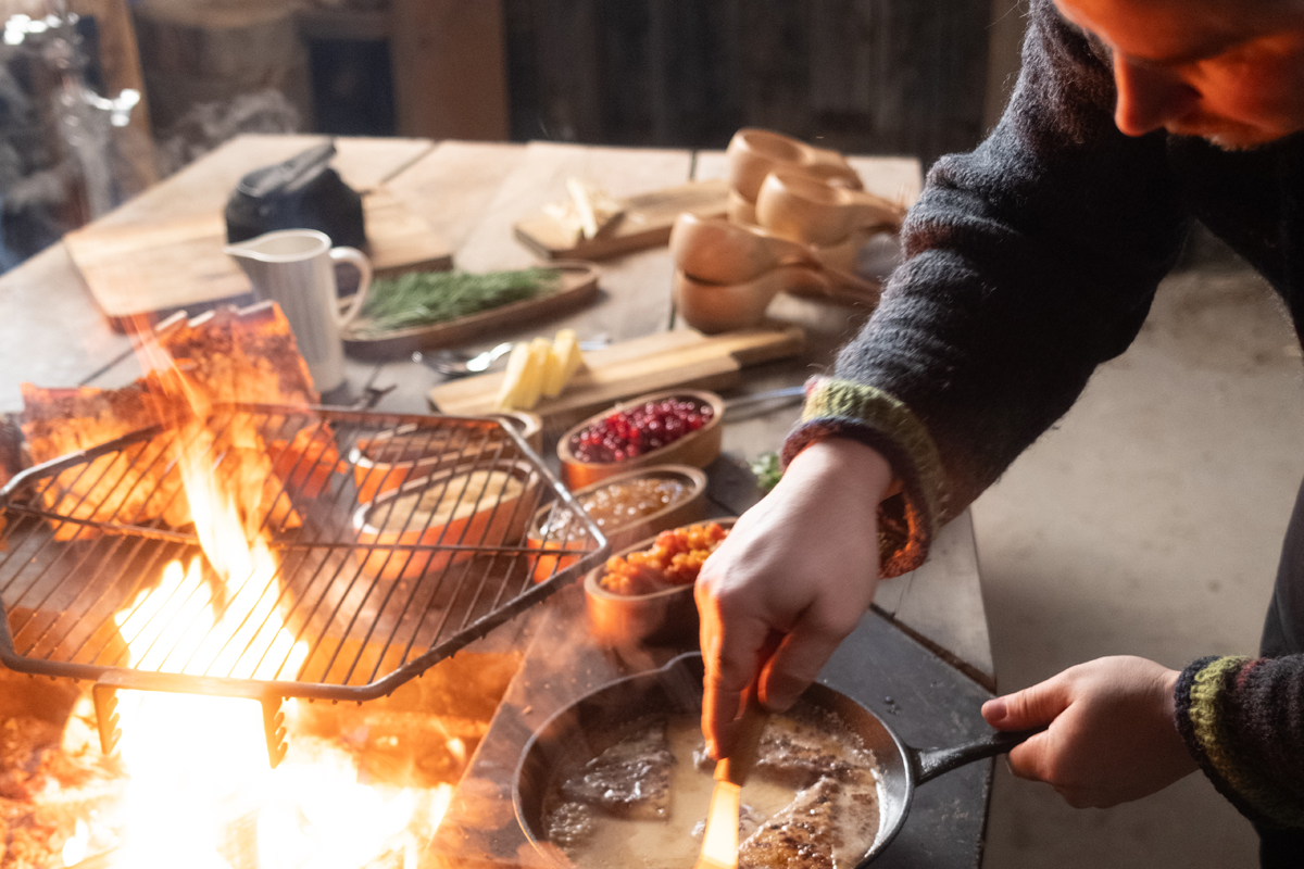Cook preparing reindeer meat in a cast iron pan over an open fire inside a kota, with wild berries and cheese, Lapland