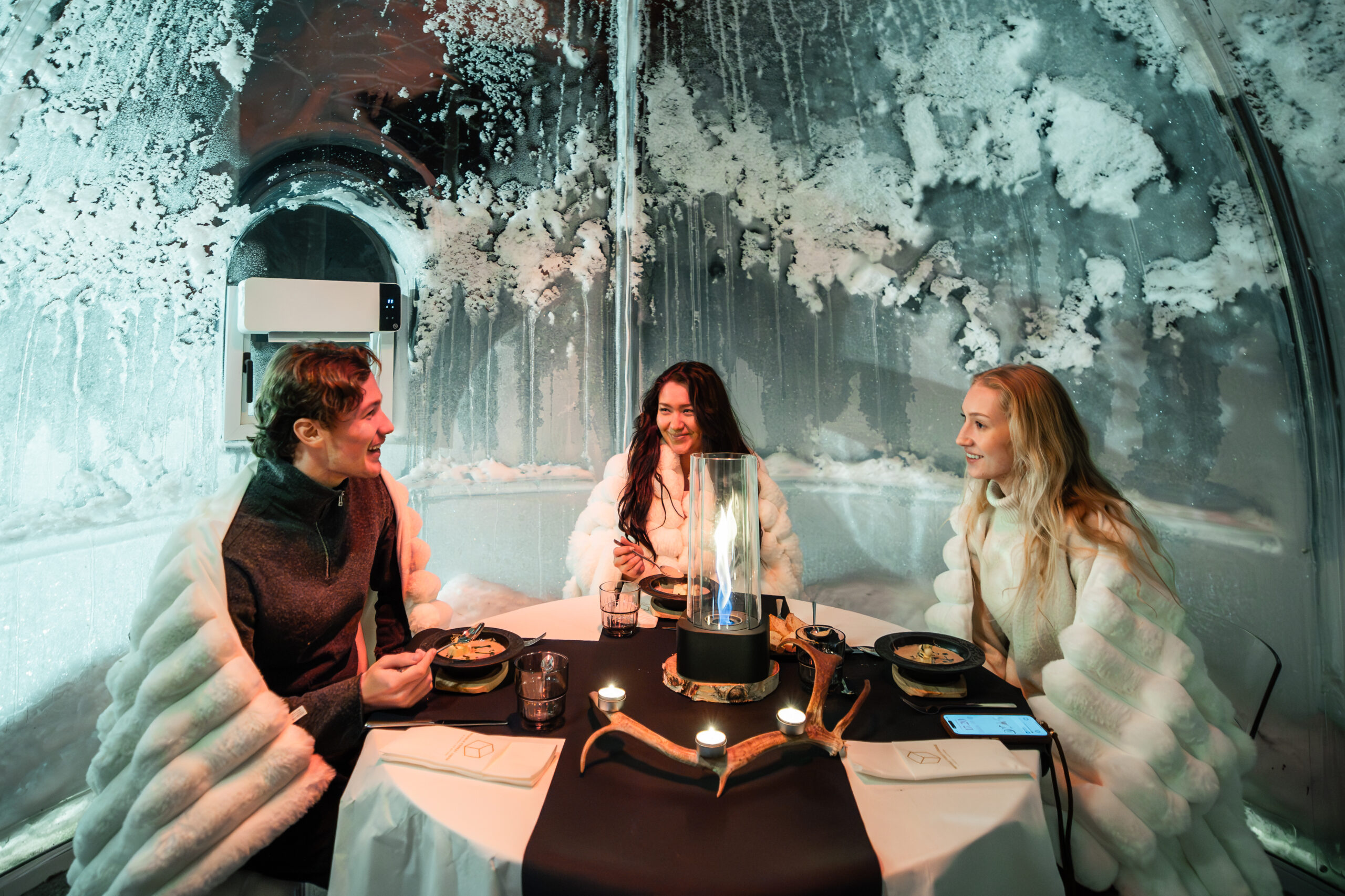 Three guests dining at a candlelit table inside a glass igloo, with snow-covered surroundings visible through the walls, Rovaniemi