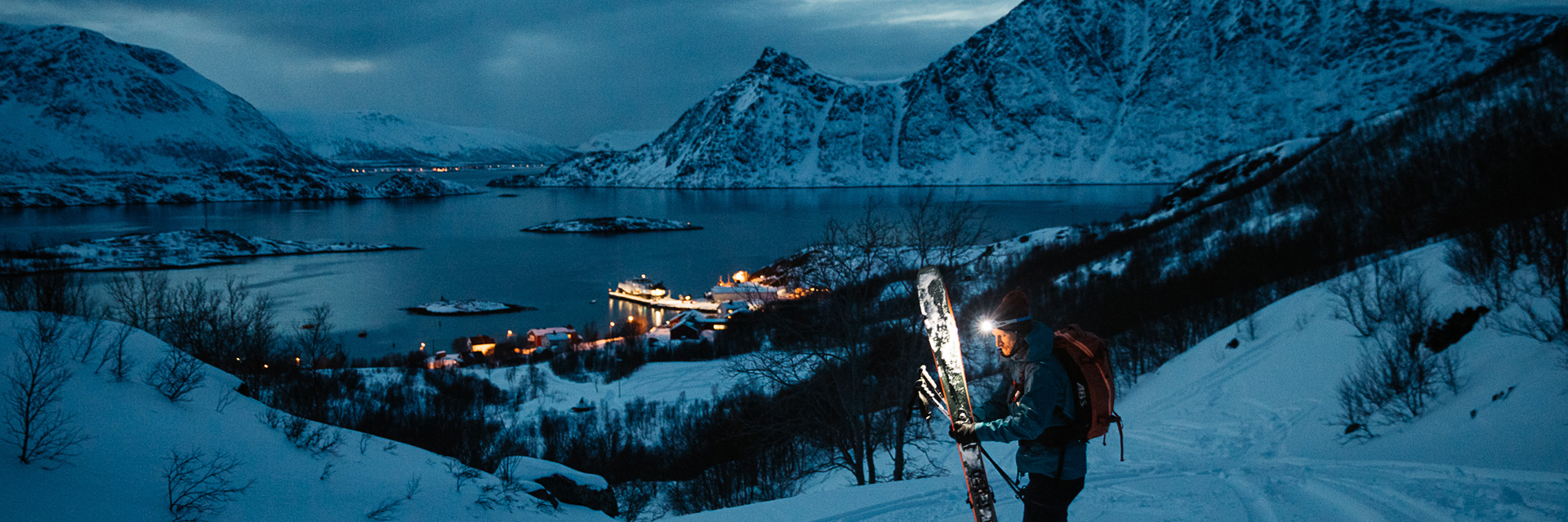 A skier with a headlamp descends a mountain above a fjord village at dusk.