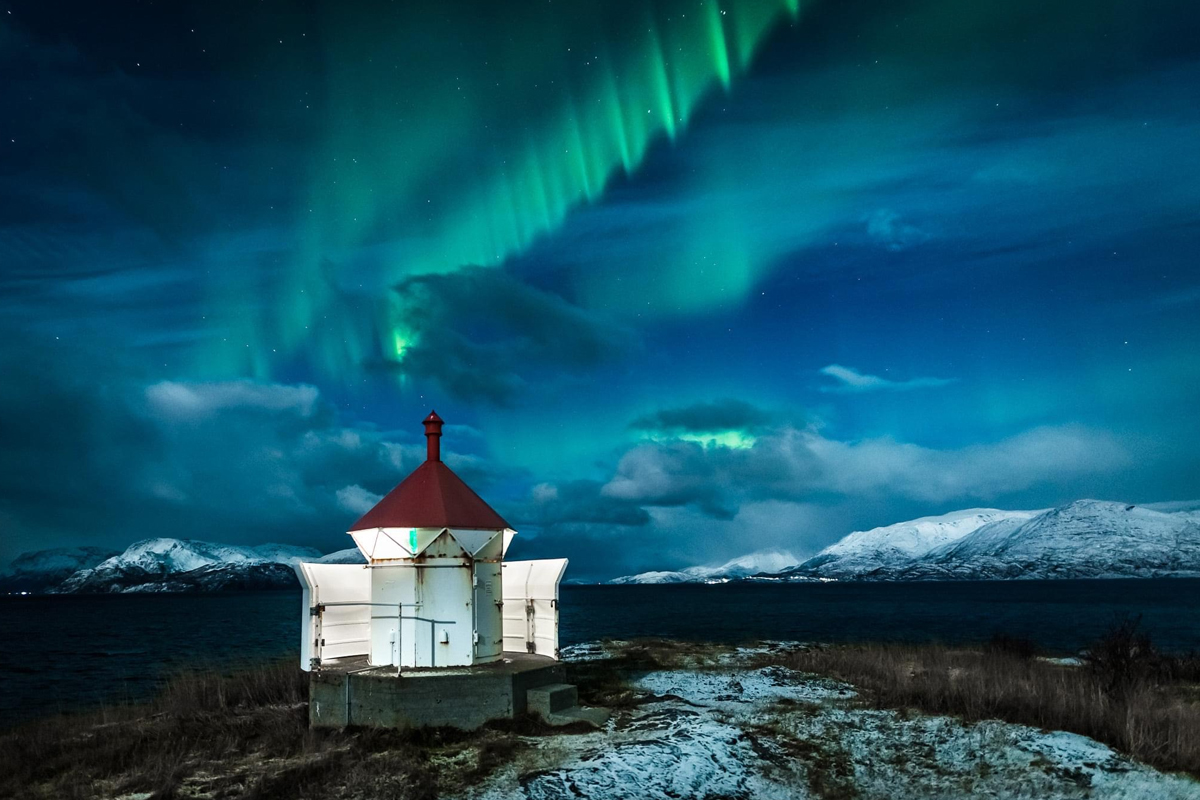 Aurora borealis glowing over a small lighthouse on the Alta coastline at night.