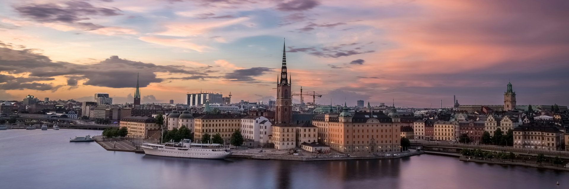 Stockholm skyline at sunset, with Riddarholmen island, church spire, and waterfront reflected in calm water.