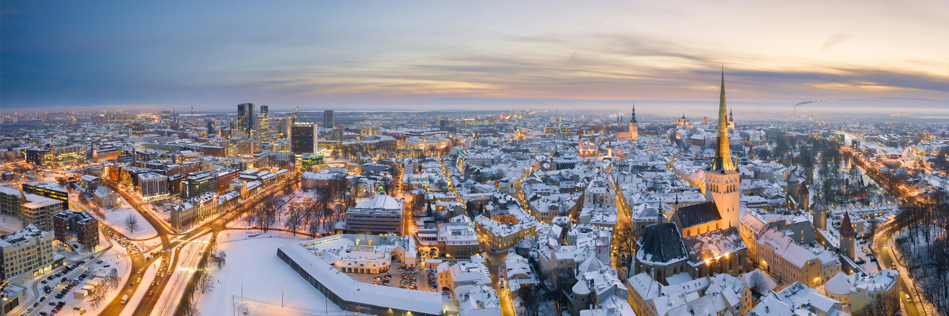 Panoramic aerial view of Tallinn at dusk in winter, showing the medieval Old Town, St Olaf's Church spire, and the modern city beyond.