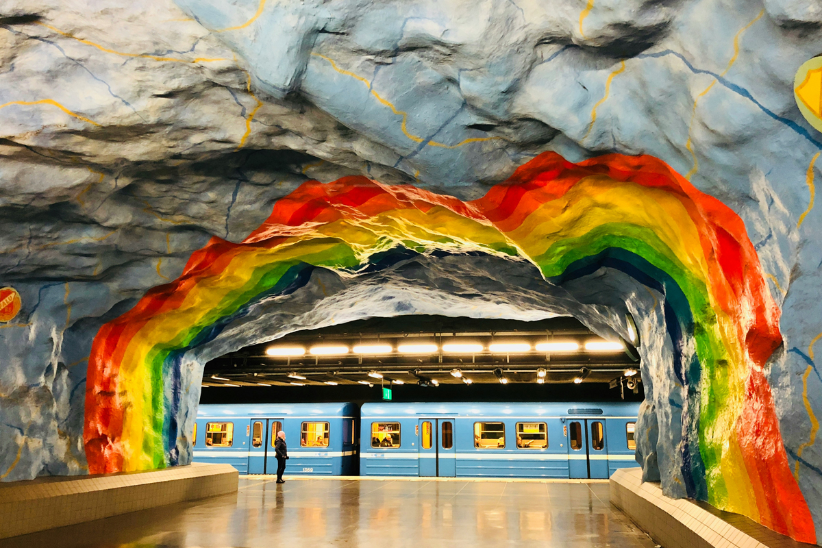 Painted rock walls and rainbow mural inside a Stockholm metro station, with a blue train on the platform.