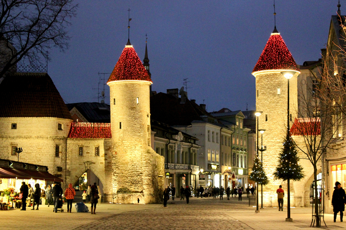 Viru Gate towers decorated with Christmas lights at dusk, with people walking through Tallinn's Old Town in winter.