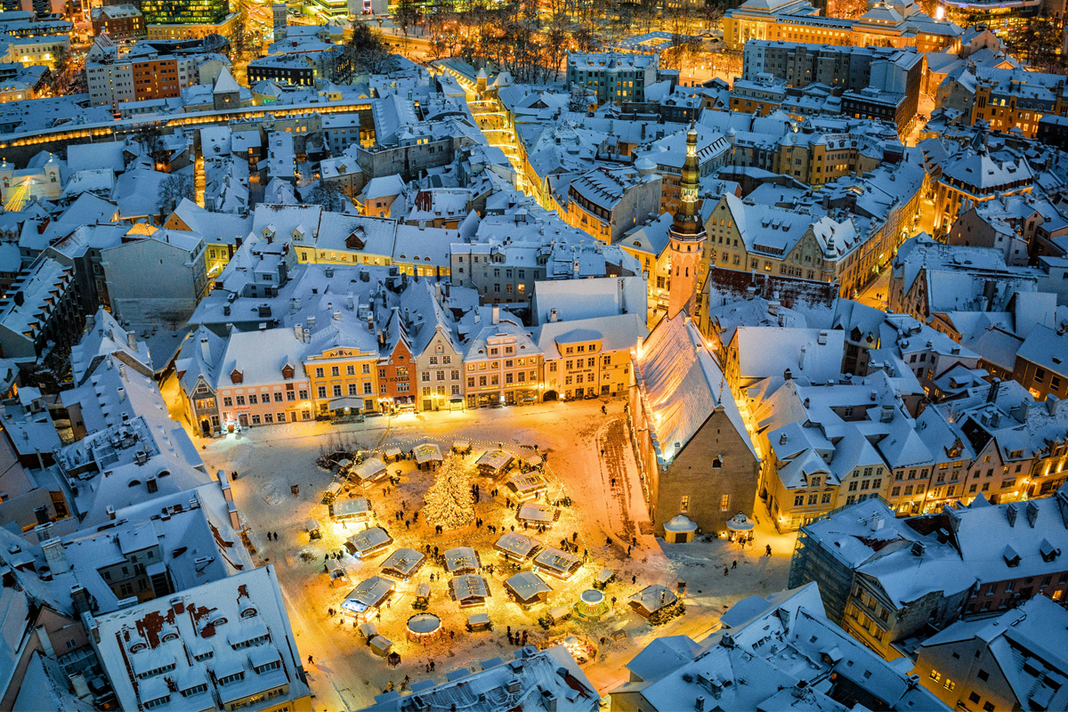 Aerial view of Tallinn's Town Hall Square at night in winter, with snow-covered rooftops and a lit Christmas market below.
