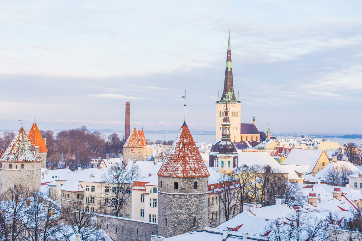 Snow-covered rooftops and medieval towers of Tallinn's Old Town in winter, with St Olaf's Church spire in the background.