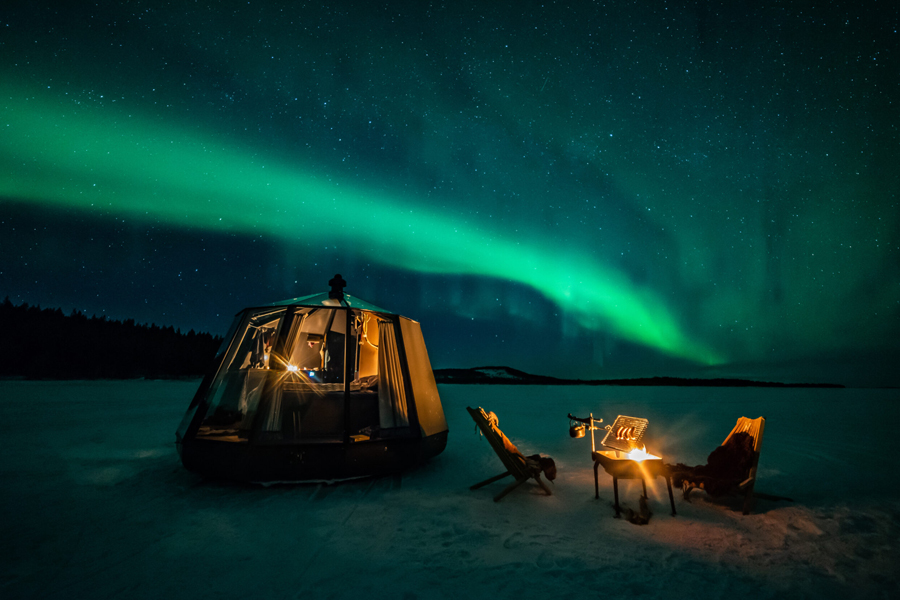 A lit glamping tent on a frozen lake with two chairs and a fire beside it under a vivid northern lights display, Jokkmokk, Swedish Lapland