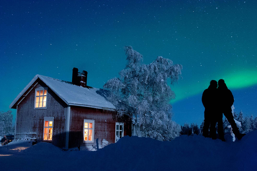 Two people silhouetted against a lit red cabin and a green northern lights display in a snowy winter landscape, Jokkmokk, Swedish Lapland. Photo: Solberg Weihnachten