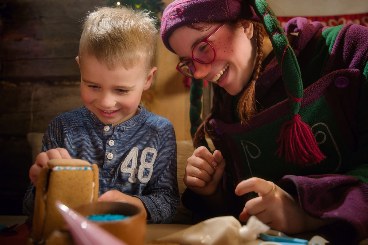 Young child decorating a gingerbread house with an elf at a Christmas workshop in Lapland.