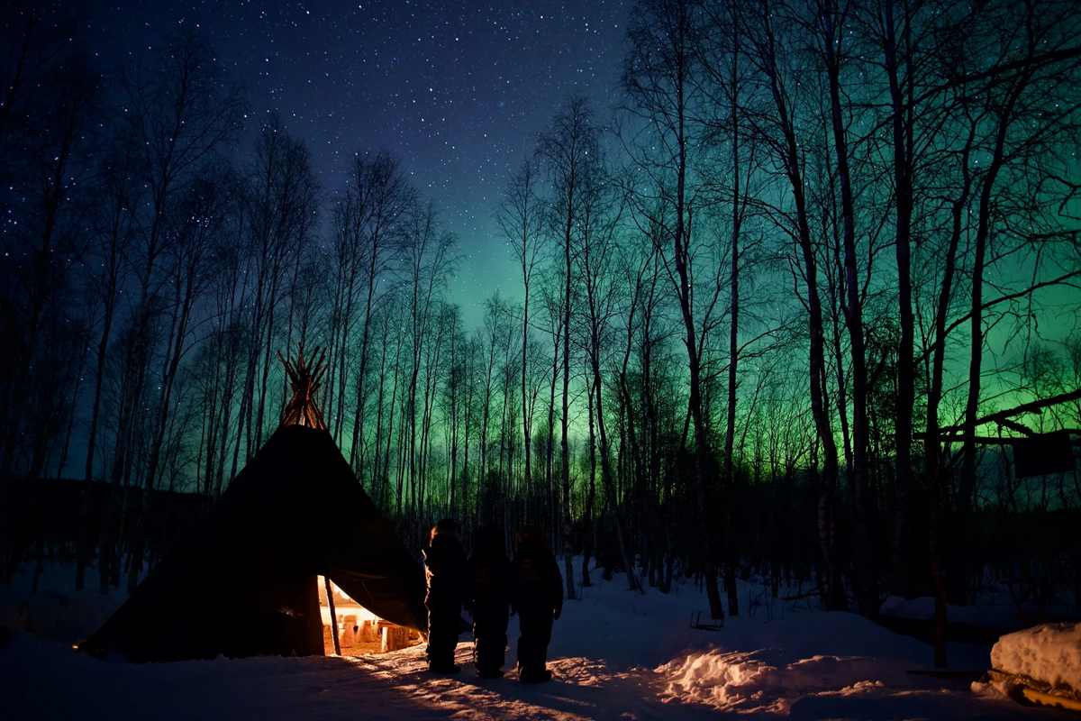 Family by a Sámi tent under the Northern Lights in a Finnish Lapland forest.