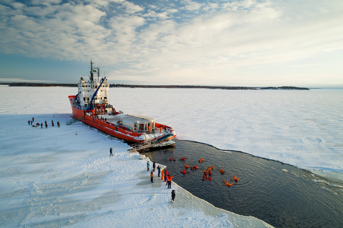 Des visiteurs en combinaison de flottaison dans l'eau libre, à côté d'un brise-glace sur la mer gelée.
