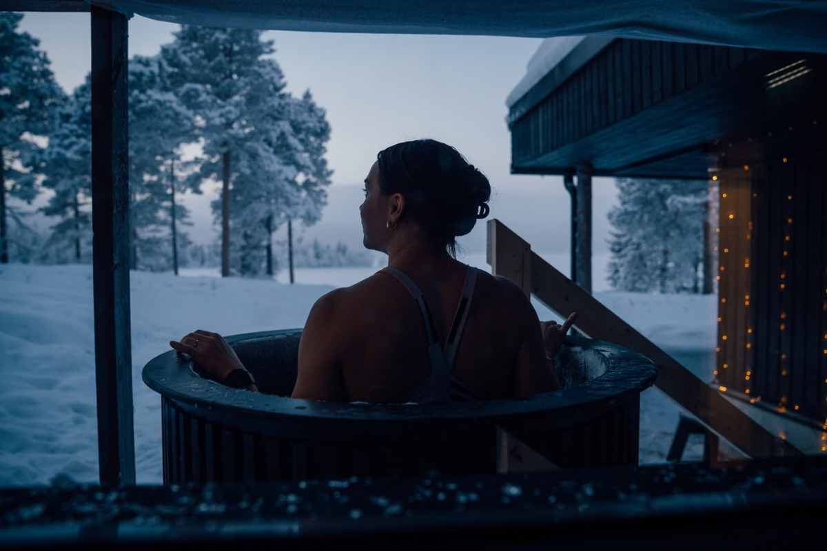 Une femme dans un bain extérieur au Kelo Resort, face à la forêt enneigée au crépuscule.