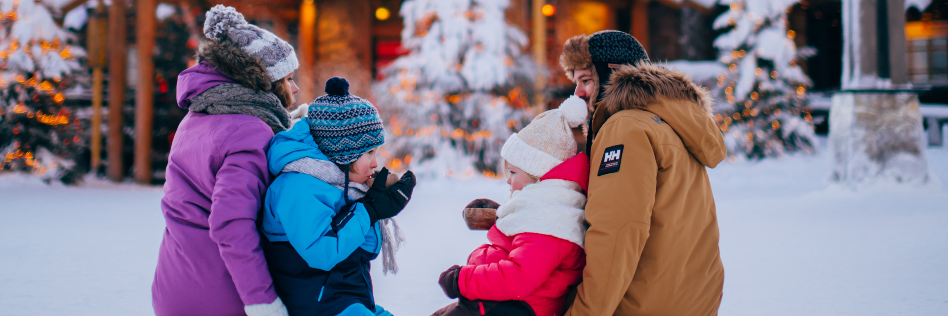 Family of four in winter clothing gathered in the snow, with Christmas-lit trees and a log building in the background, Rovaniemi