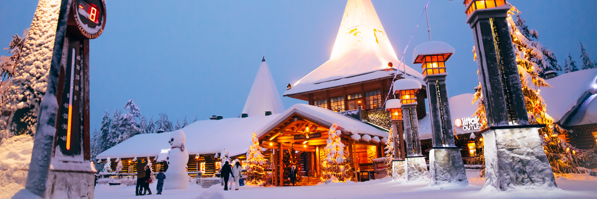 Santa Claus Village at dusk, snow-covered log buildings lit with Christmas lights and families walking in the square, Rovaniemi