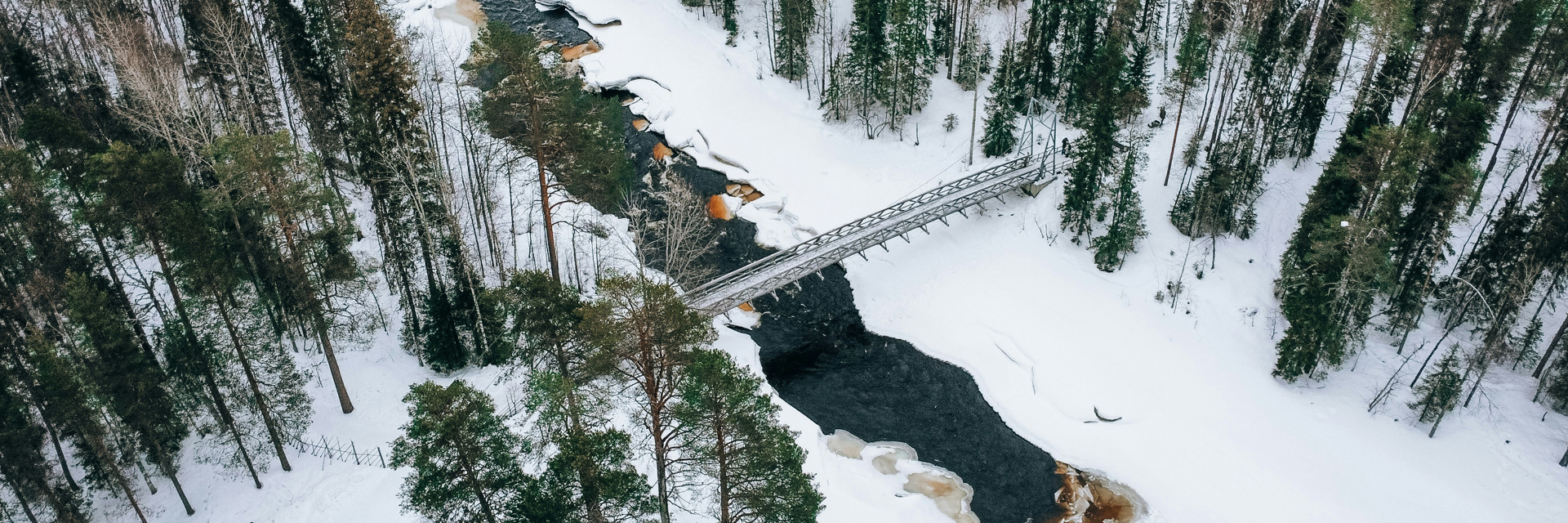 Aerial view of a footbridge crossing a partly frozen river through a snowy pine forest in Lapland.