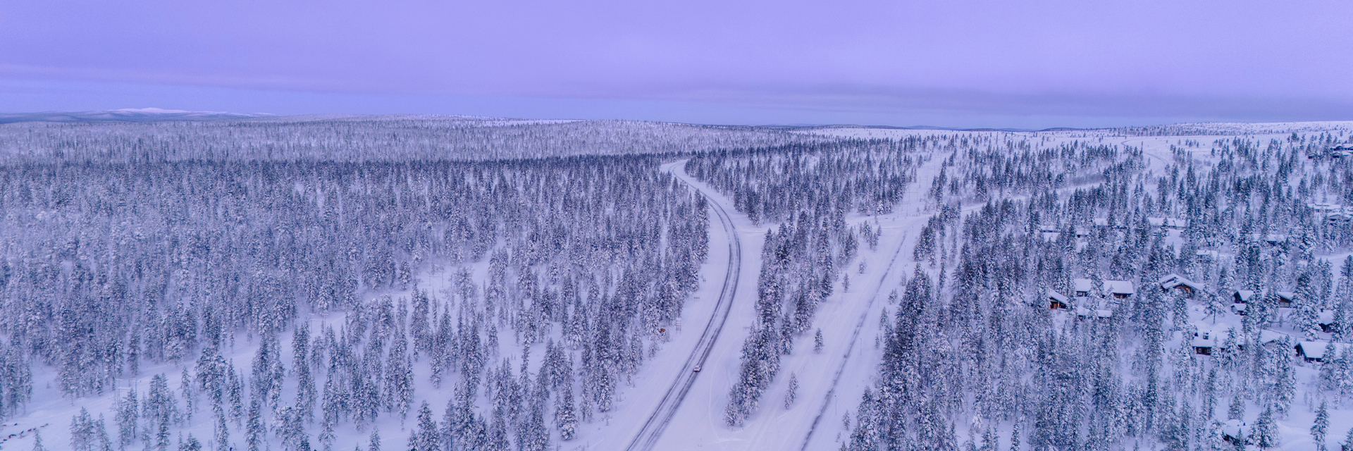 Aerial view of a snow-covered road winding through a vast pine forest under a purple Arctic sky.
