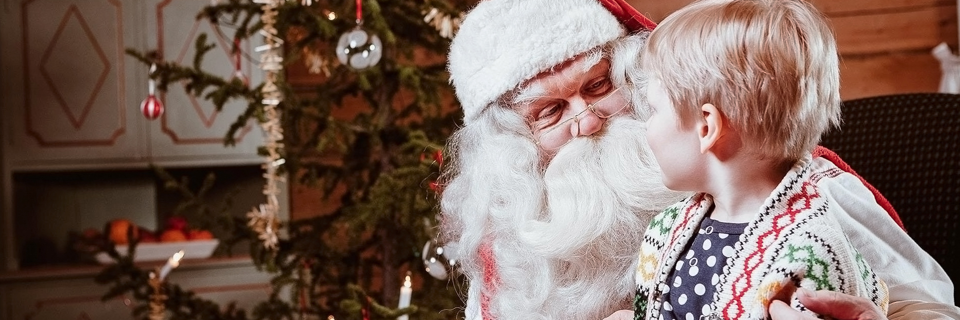 Santa Claus shares a warm moment with a young child by the Christmas tree in a cozy Rovaniemi cabin.