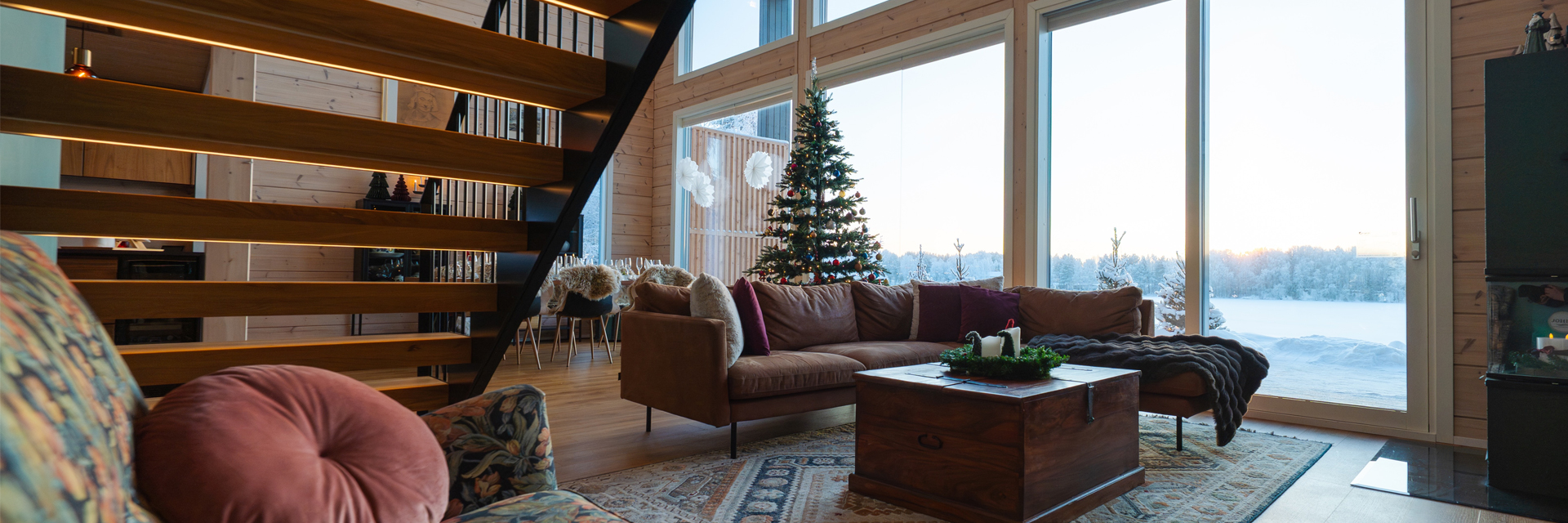 A Christmas tree and cozy sofas fill a wood-lined living room with snowy lake views in Rovaniemi.