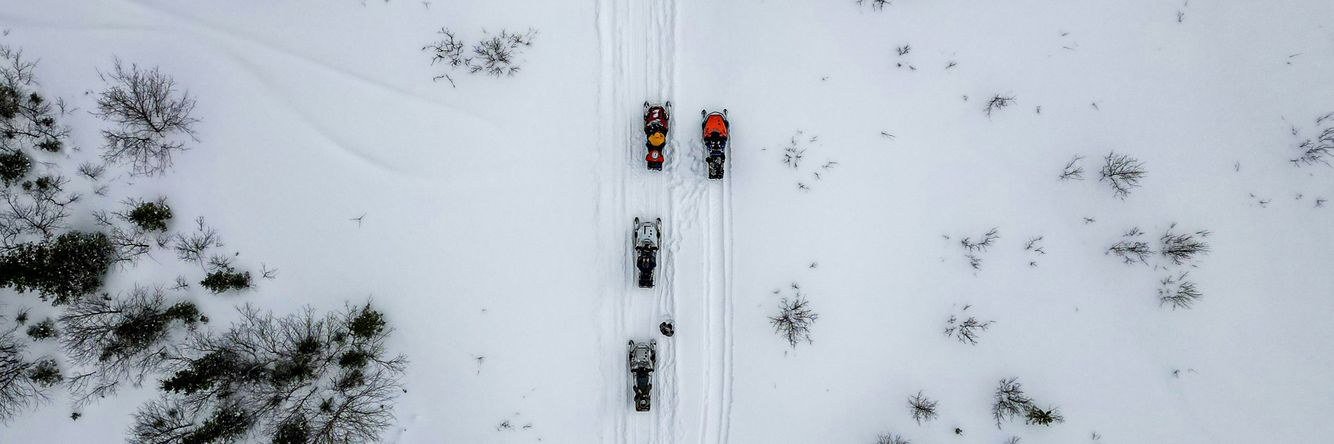 Aerial view from directly above four snowmobiles riding in a line across an open snowfield.