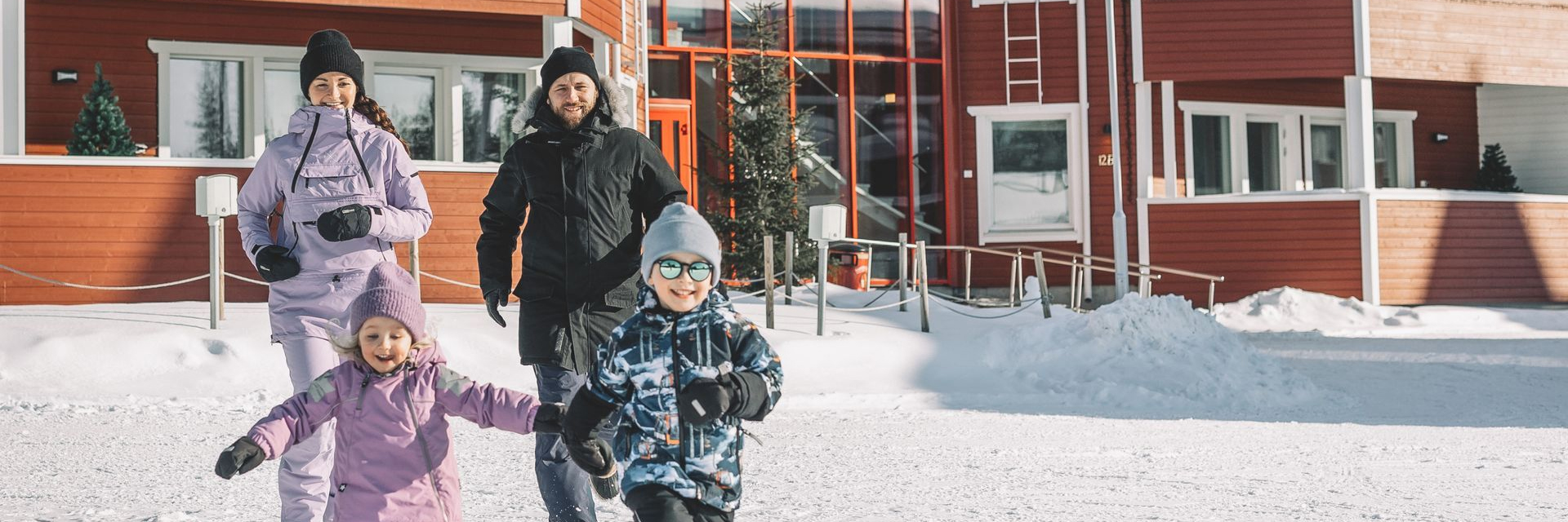 Family of four running and laughing in the snow outside a red building at Santa Claus Holiday Village, Rovaniemi