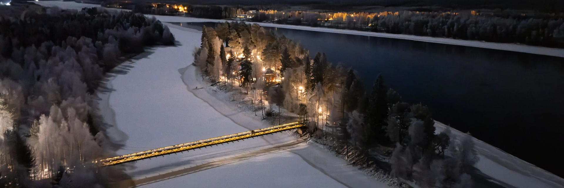 Aerial night view of Aino Private Island with a lit footbridge, illuminated buildings and frost-covered trees on the frozen Kemijoki river, Rovaniemi