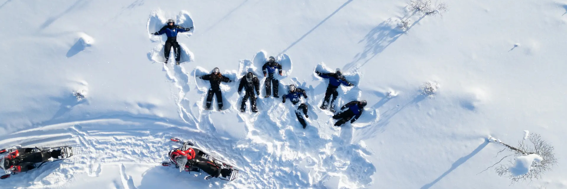 Aerial view of a group making snow angels in snowmobile suits on a frozen lake, with snowmobiles parked nearby, Kemijärvi