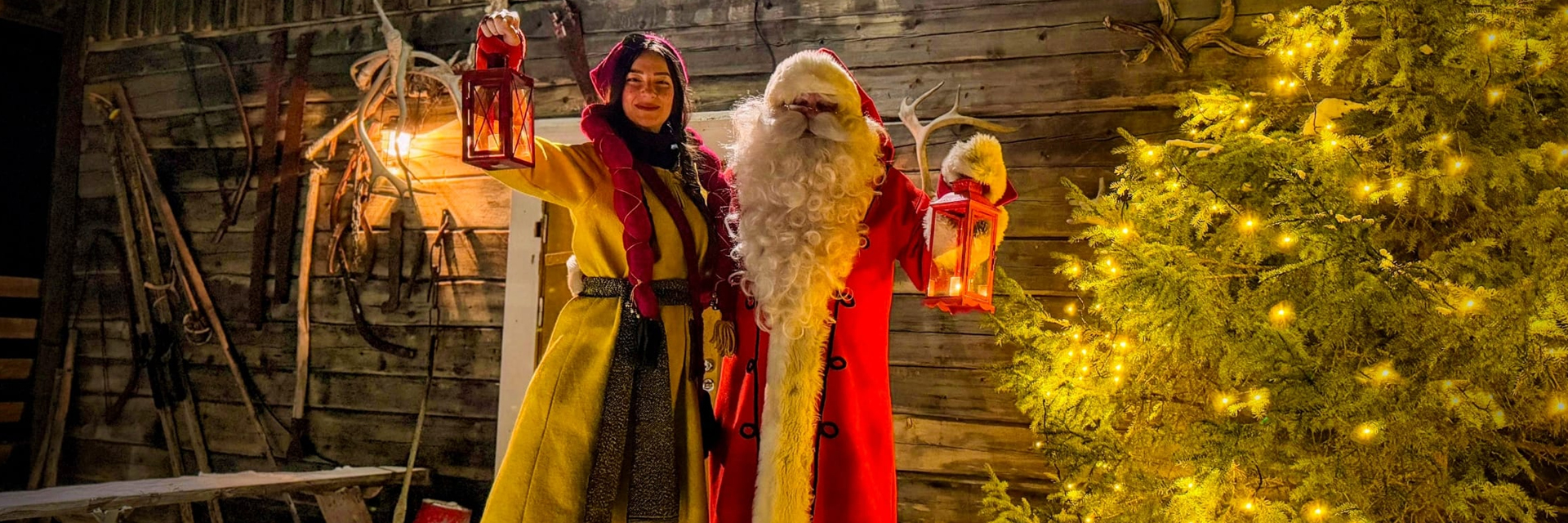 Santa Claus and an elf in traditional costumes holding lanterns beside a lit Christmas tree, Levi, Finnish Lapland