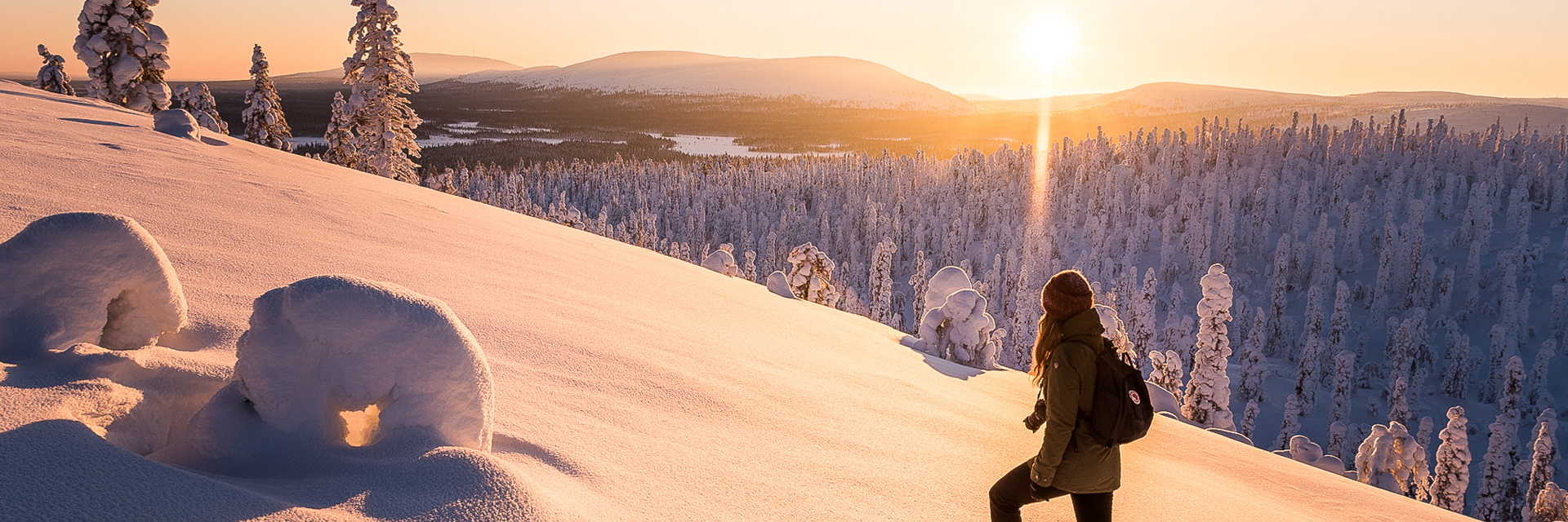 A person walks across a snow-covered fell slope at sunset, with frost-laden trees and open winter landscape stretching into the distance in Ylläs, Finland.